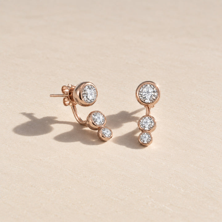 Rose gold three-stone moissanite ear jacket earrings with graduated round stones. displayed on a light beige textured surface.