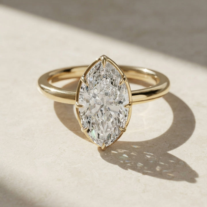 A yellow gold ring featuring a 3 carat marquise-cut clear moissanite in a prong setting, resting on a textured beige surface with soft shadows.