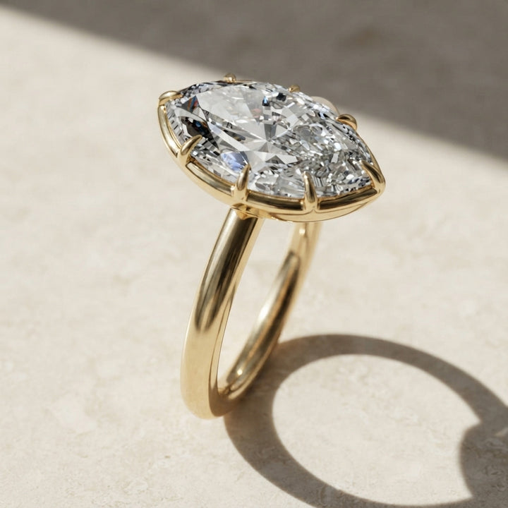 A yellow gold ring featuring a large marquise-cut clear moissanite in a prong setting, resting on a light beige textured surface with a soft shadow.
