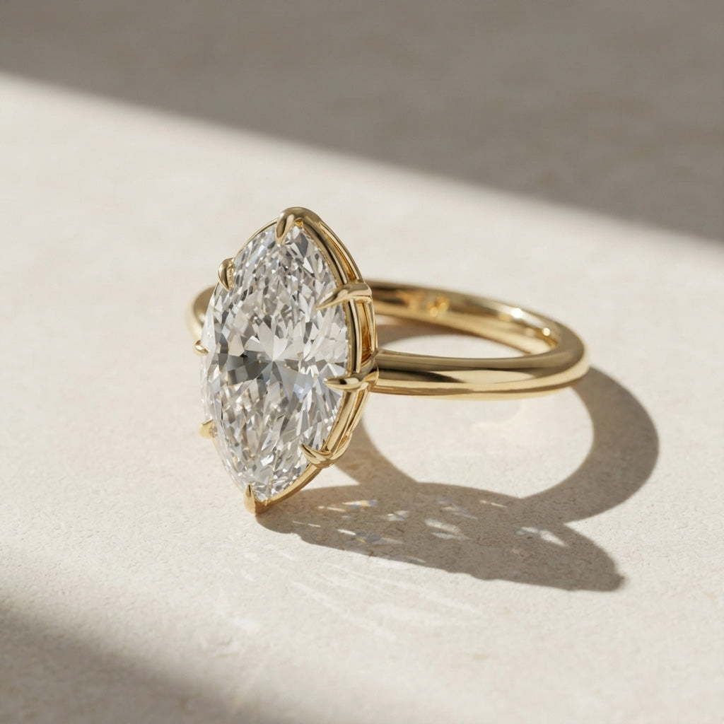 A yellow gold ring featuring a large, brilliant-cut diamond in a marquise-shaped bezel setting, resting on a textured beige surface with dramatic diagonal shadows.