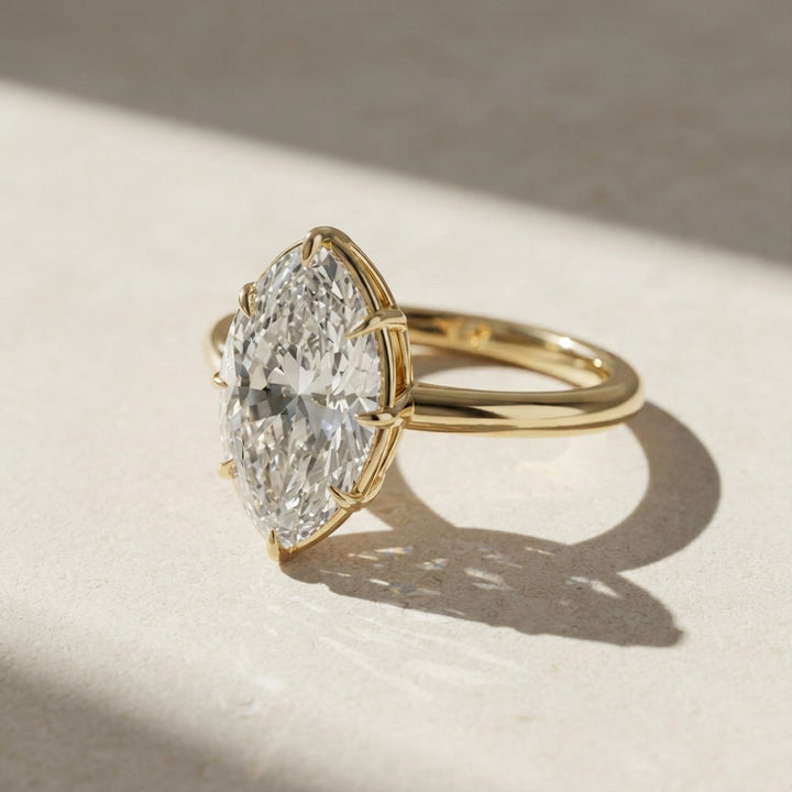 A yellow gold ring featuring a large, brilliant-cut diamond in a marquise-shaped bezel setting, resting on a textured beige surface with dramatic diagonal shadows.