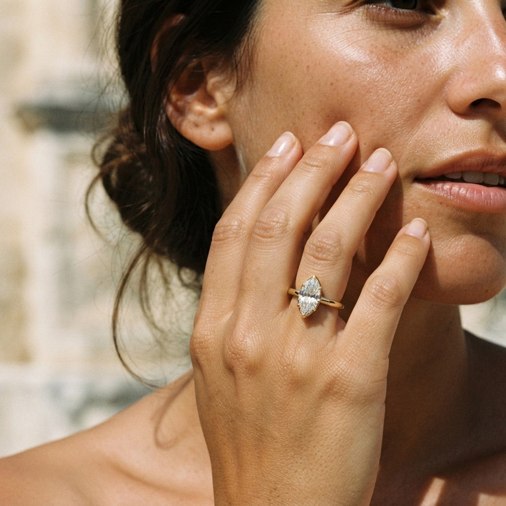 A close-up lifestyle shot of a woman wearing a gold ring with a marquise-cut clear moissanite on her finger, with her hand resting on her cheek against a softly blurred outdoor background.