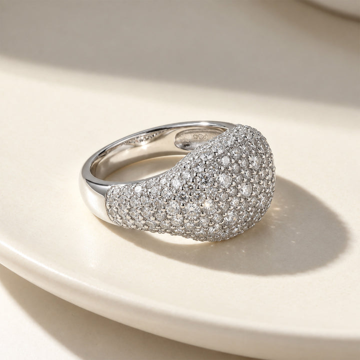 A silver pavé dome moissanite ring, resting on a warm cream ceramic surface with soft shadows.
