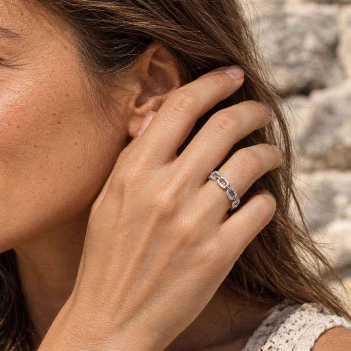 A close-up lifestyle shot of a woman wearing a delicate pavé chain-link moissanite ring on her ring finger, set against a soft-focus stone wall background.
