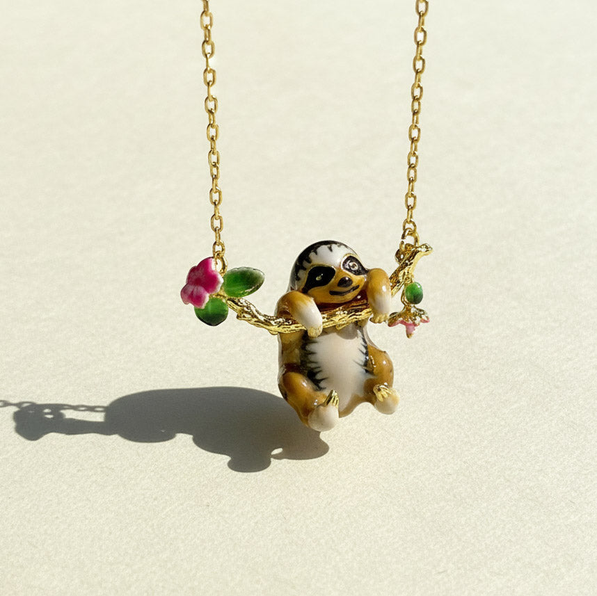 Gold chain necklace featuring a hand-painted enamel sloth pendant hanging from a floral branch with pink flowers and green leaves
