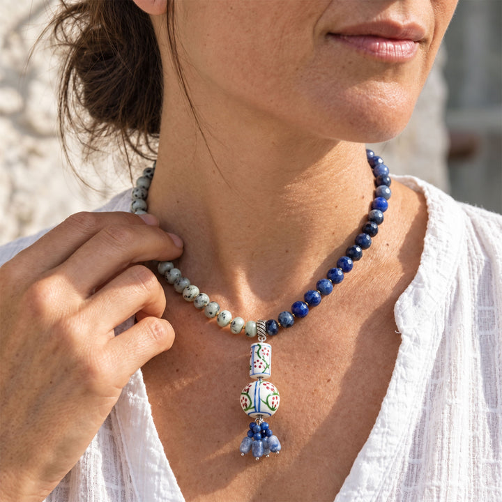 A close-up lifestyle shot of a woman wearing a beaded necklace. The necklace features a strand of dark blue lapis lazuli and a contrasting strand of light blue and white marbled beads, accented by a colorful hand-painted ceramic pendant with blue tassels.