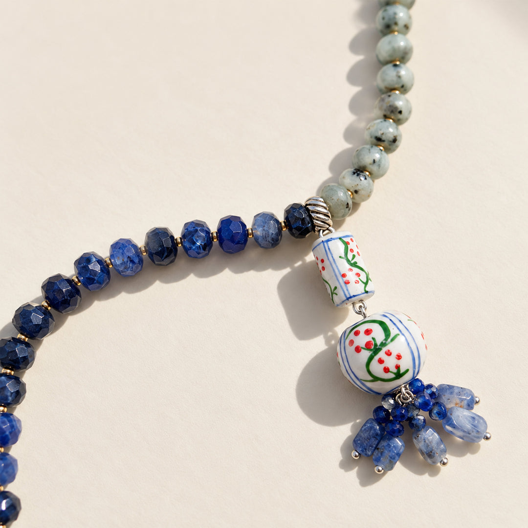 A beaded necklace featuring faceted deep blue lapis beads, light blue-green stone beads, and a decorative hanging charm with hand-painted white ceramic-style details and blue bead tassels, shown on a warm off-white background.