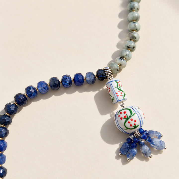 A beaded necklace featuring faceted deep blue lapis beads, light blue-green stone beads, and a decorative hanging charm with hand-painted white ceramic-style details and blue bead tassels, shown on a warm off-white background.