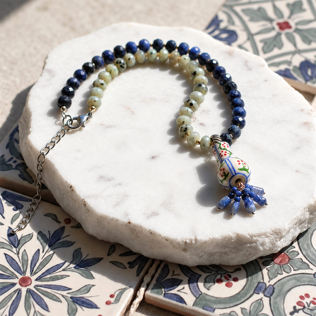A beaded necklace with lapis beads and pale green beads, a silver clasp, and a colorful ceramic-style pendant, displayed on a white stone slab over a floral tile surface.