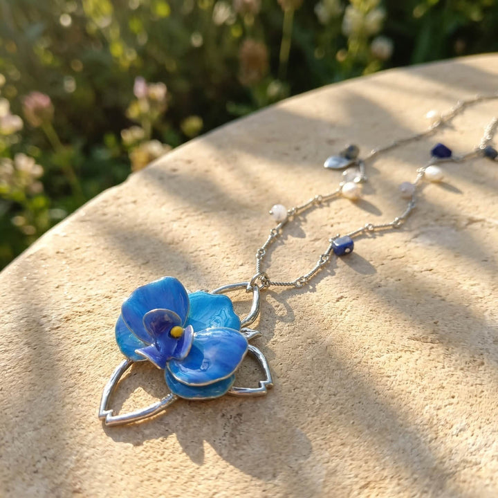 Blue flower pendant necklace on a stone surface with greenery in the background