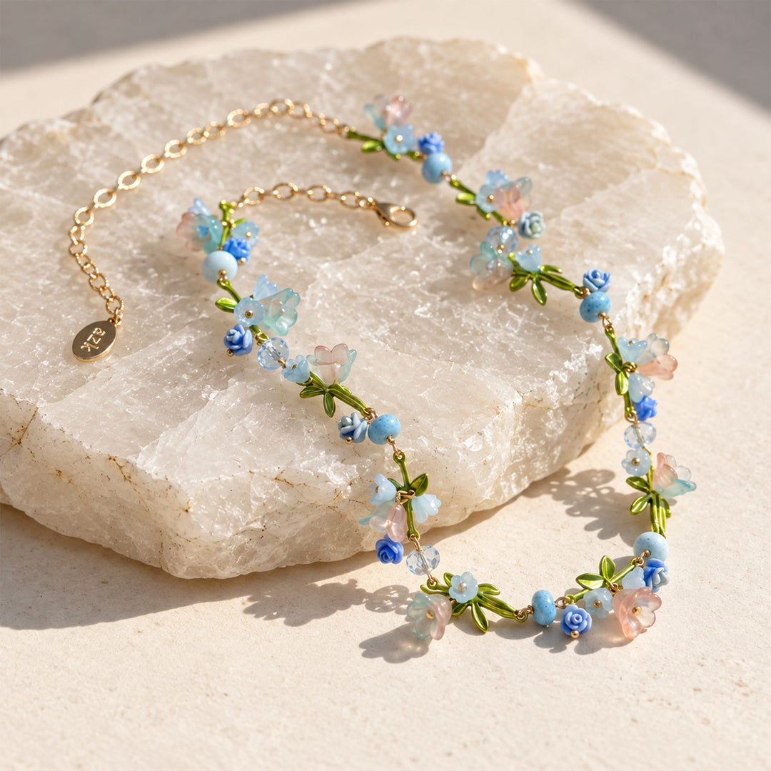 Pastel floral necklace with glass flowers, resin blooms and green enamel leaves displayed on a sunlit stone surface.