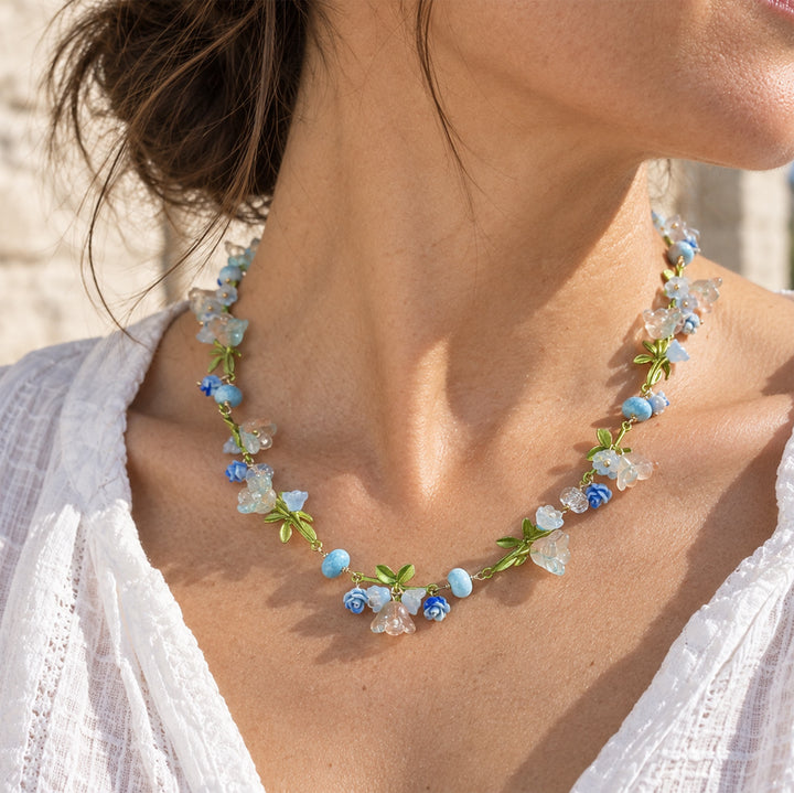 Model wearing a pastel floral necklace with glass flowers and enamel leaves outdoors in natural sunlight.