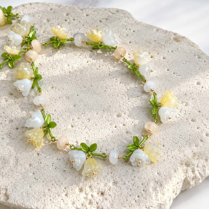 Close-Up of Handcrafted Floral Necklace with Glass Bellflowers and Green Enamel Leaves on a textured stone surface