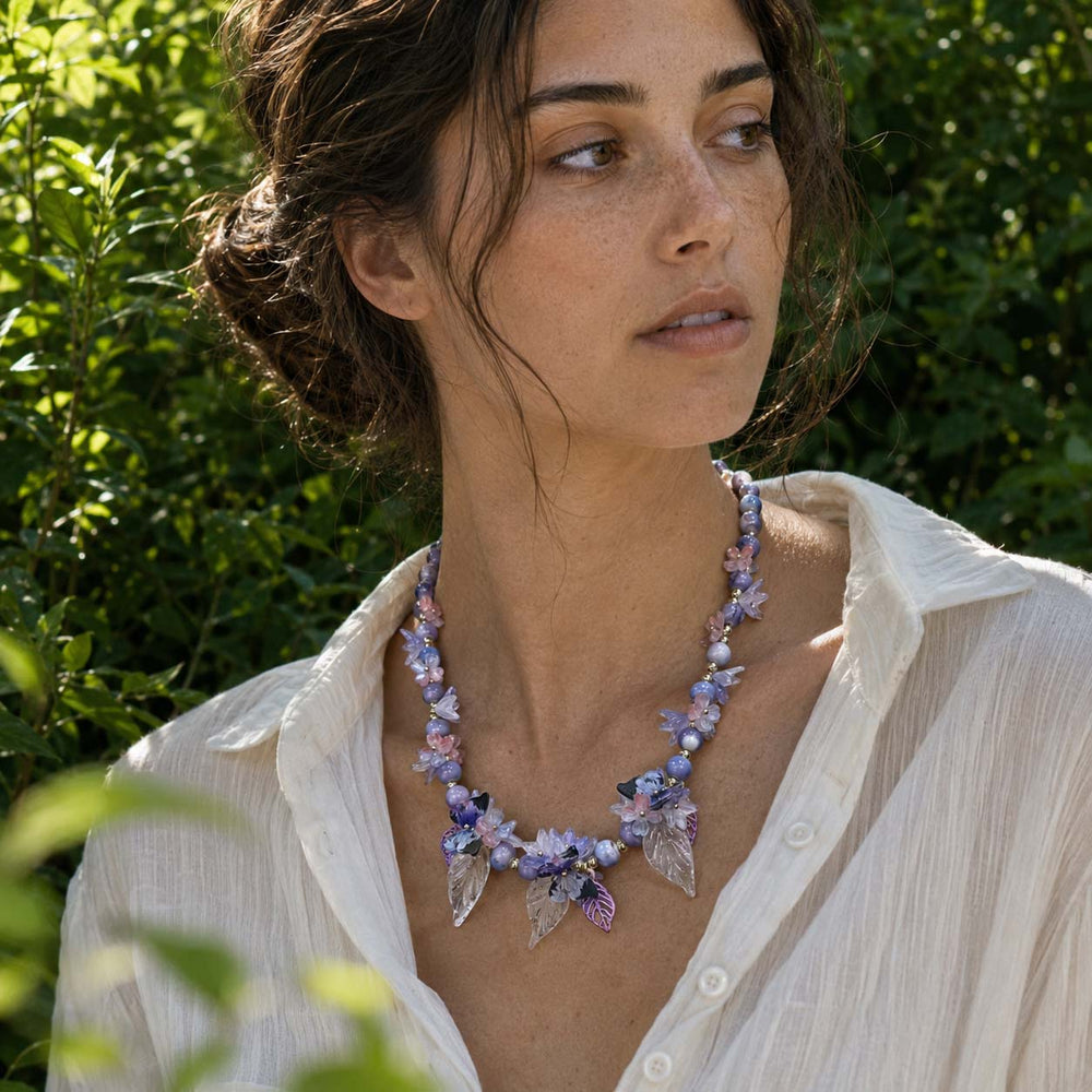 Woman wearing a Butterfly Bloom Garden Necklace with Lavender Glass Flowers and Resin Butterfly Details outdoors with greenery in the background