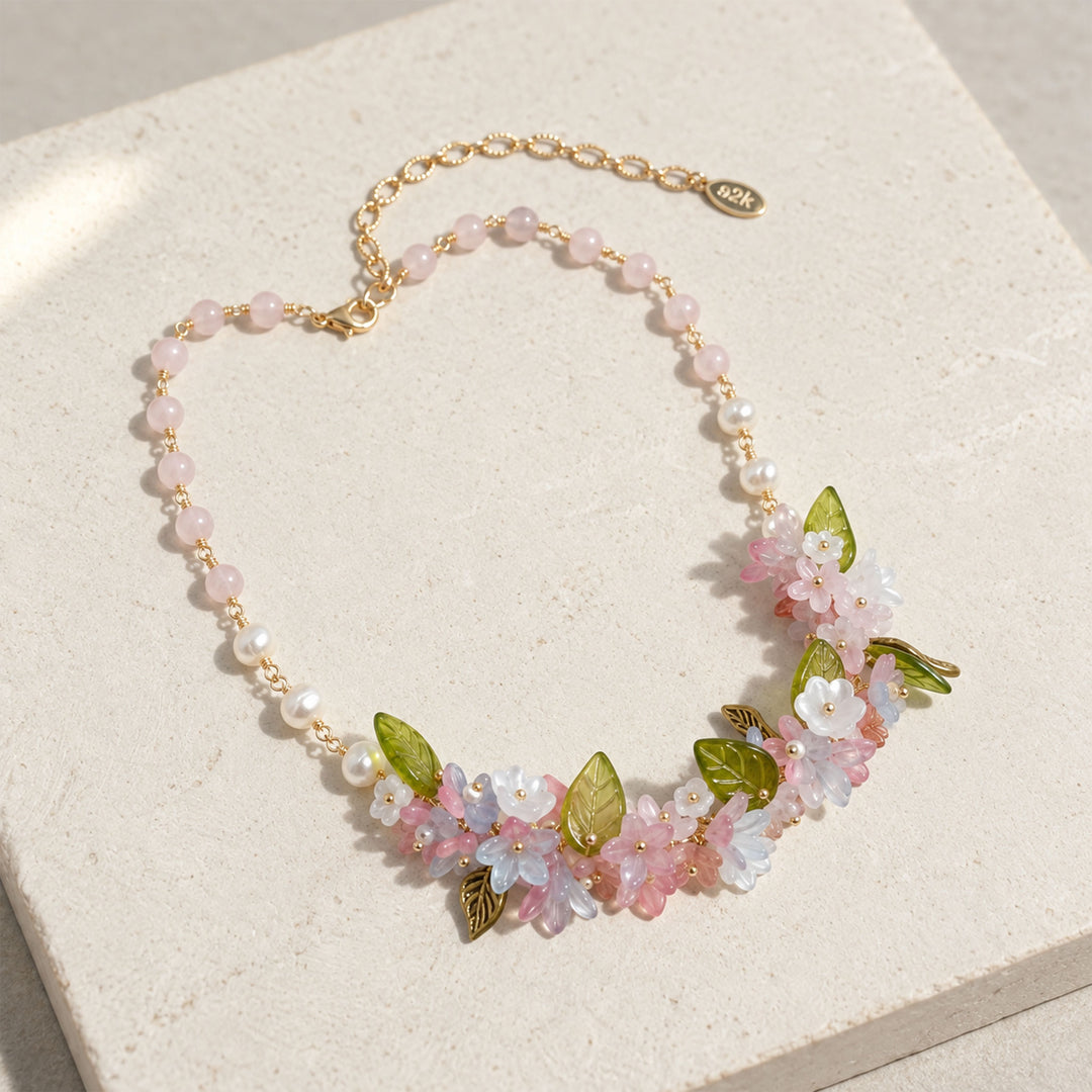 Pink quartz floral necklace with freshwater pearls, glass and resin flowers, and green glass leaves on pale stone, displayed on a textured cream stone block.