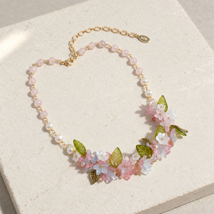 Pink quartz floral necklace with freshwater pearls, glass and resin flowers, and green glass leaves on pale stone, displayed on a textured cream stone block.