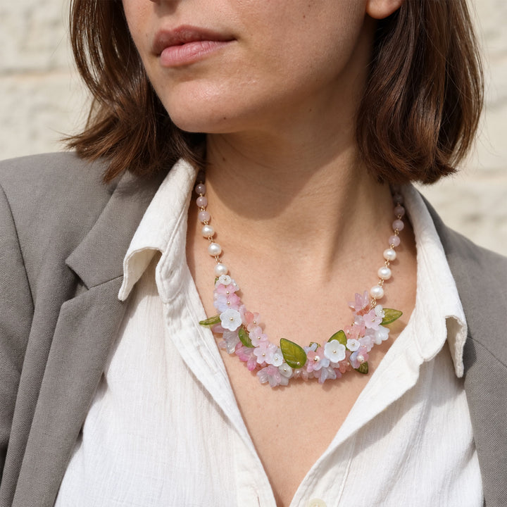 A close-up lifestyle shot of a woman wearing a delicate floral necklace with pink and white flowers, green glass leaves, and freshwater pearls and pink quartz beads, styled with a white shirt and gray blazer.