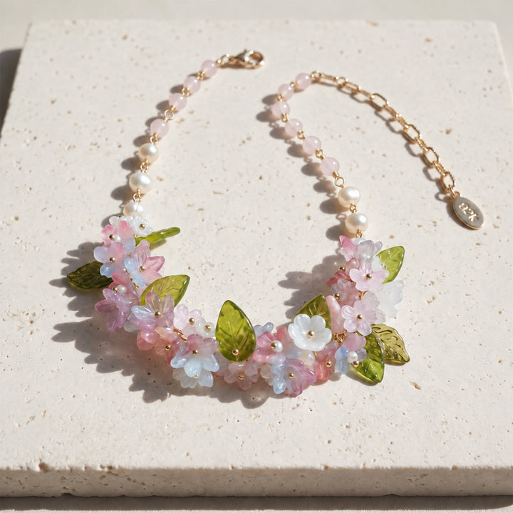 A delicate statement necklace featuring pink and white glass flower beads, translucent green leaves, freshwater pearls and pink quartz beads, laid out on a textured beige stone tile.