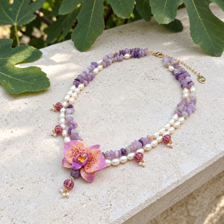 A double-strand beaded necklace featuring freshwater pearls, lavender and purple amethyst chips, pink faceted lampwork beads, and a central pink orchid flower charm, displayed on a light stone surface with green leaves in the background.
