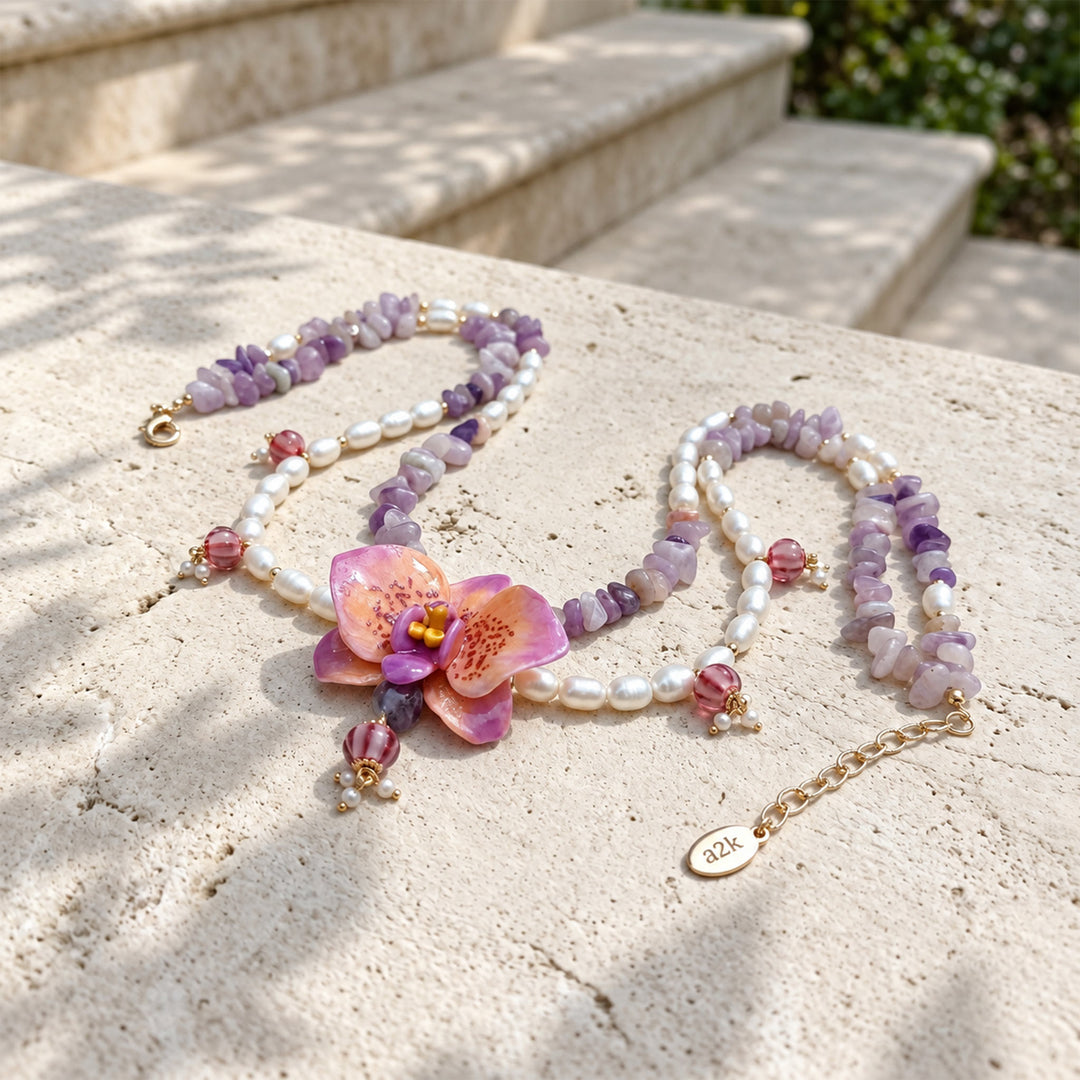 A beaded necklace with freshwater pearls, purple jade chips, pink faceted lampwork glass beads, and a central pink orchid flower charm, displayed on a light stone surface outdoors.