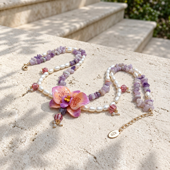 A beaded necklace with freshwater pearls, purple jade chips, pink faceted lampwork glass beads, and a central pink orchid flower charm, displayed on a light stone surface outdoors.