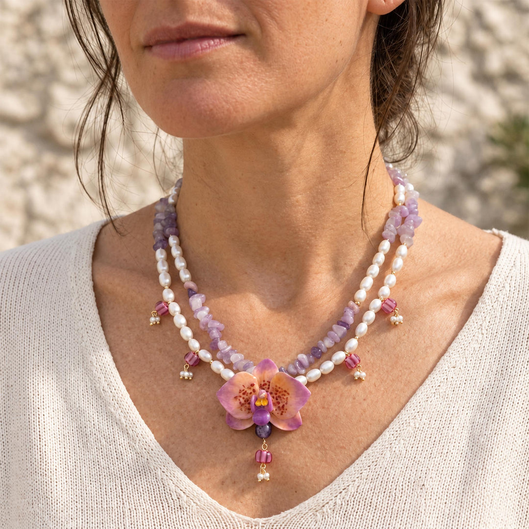 A close-up of a woman wearing a double-strand beaded necklace with freshwater pearls, purple jade beads, and a central hand painted purple orchid flower pendant.