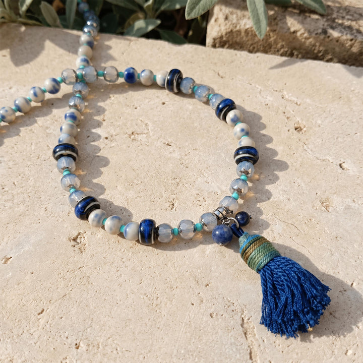 Close-up of a handcrafted lapis tassel necklace with hand-painted porcelain and faceted glass beads on pale stone.