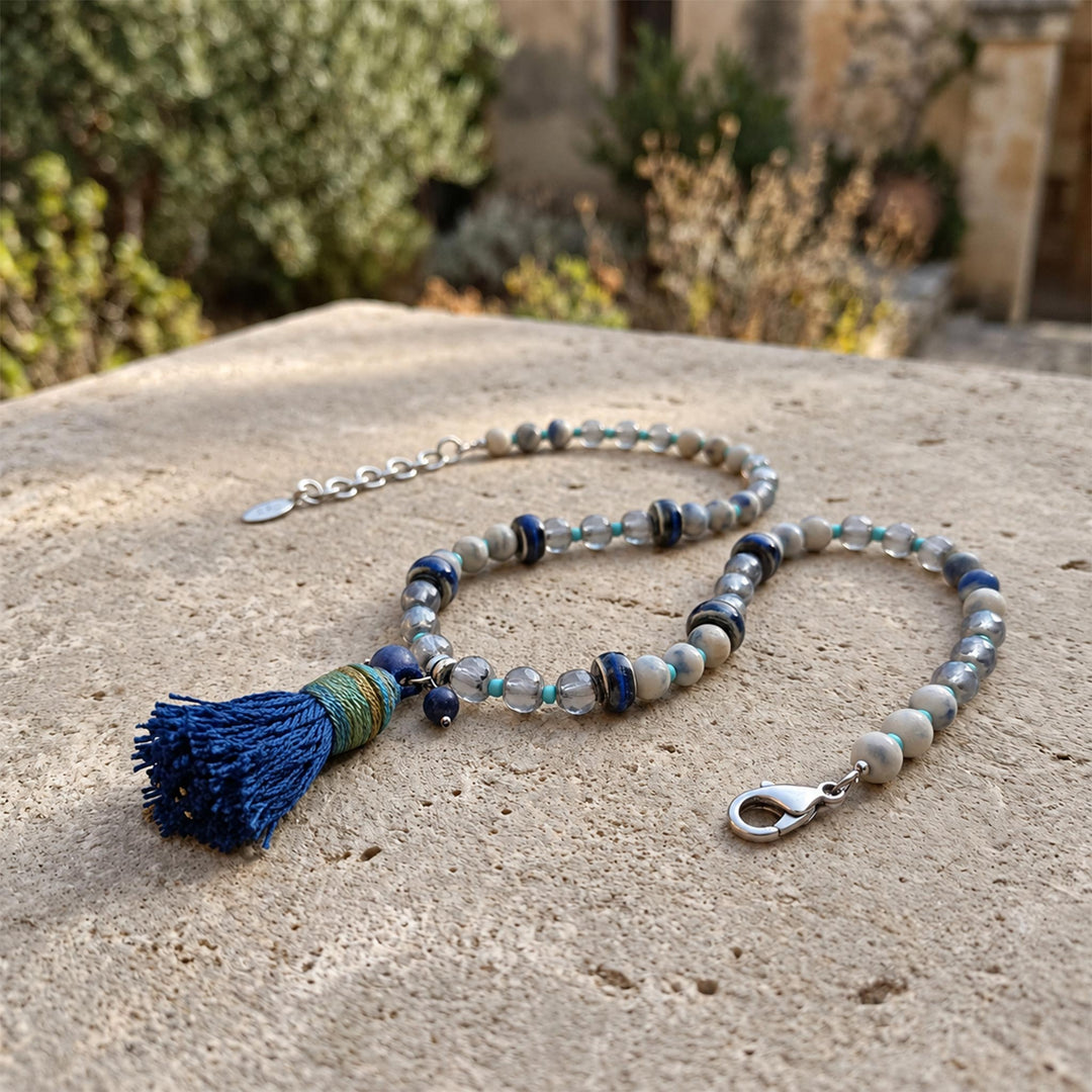 Lapis and porcelain tassel necklace with blue glass beads displayed on a sunlit pale stone surface outdoors.