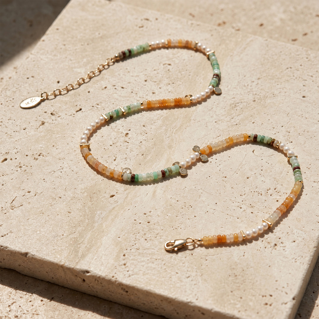 A beaded chain necklace with Chrysoprase, Yellow Jade and freshwater pearl necklace with gold disc charms displayed on a beige stone surface.