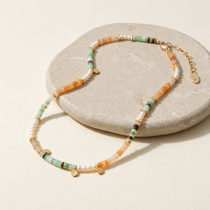 Full mixed gemstone necklace with green Chrysoprase, Yellow Jade, freshwater pearls and gold extender chain displayed on a round stone prop against a warm cream background.