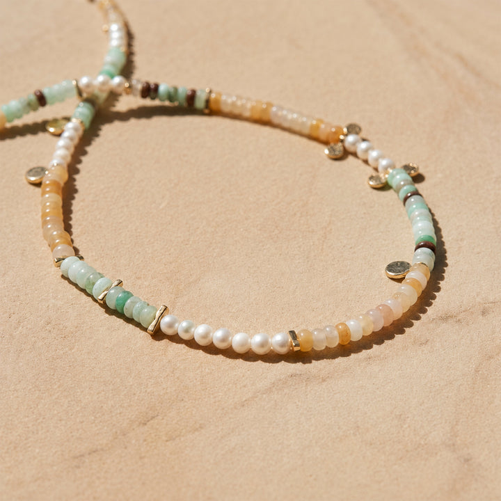 A delicate beaded strand necklace with Chrysoprase, Yellow Jade and freshwater pearl necklace with gold disc charms, arranged on a sandy beige background.