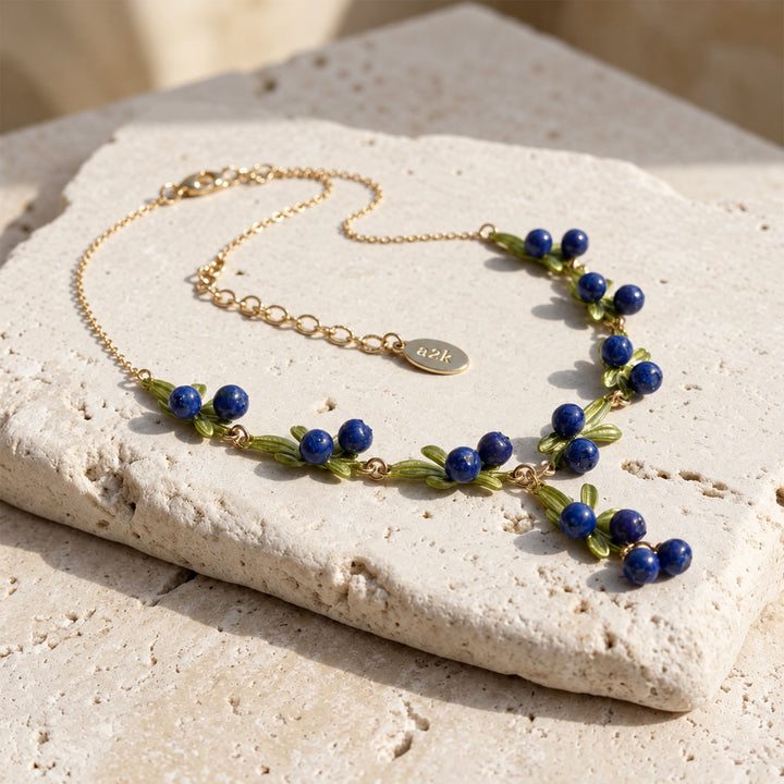 Hand-painted blueberry enamel necklace with green leaves displayed on a sunlit pale stone surface.