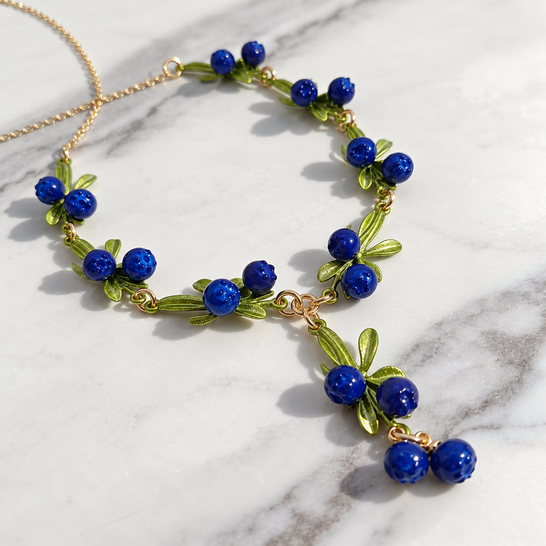 Blueberry botanical necklace with blue enamel berries and sculptural green leaves on a marble background.