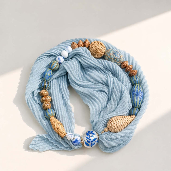 Artisan pleated scarf necklace with navy printed scarf, porcelain beads, hand-woven rattan beads and pearl magnetic clasp on a neutral background.