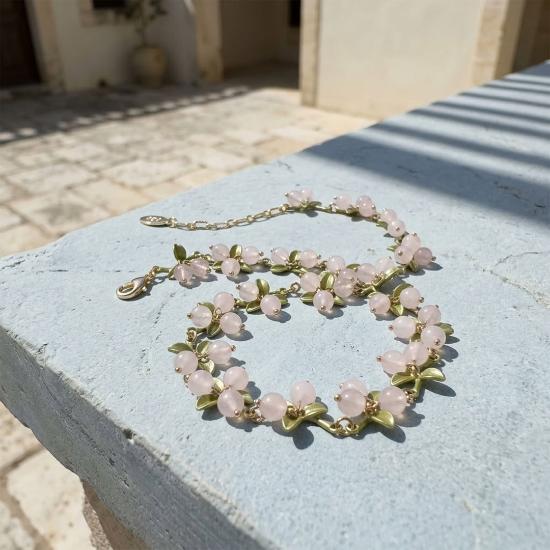 Rose Quartz Berry Garden Necklace with hand-painted green enamel leaves and gold-plated details displayed on a blue outdoor surface in sunlight.