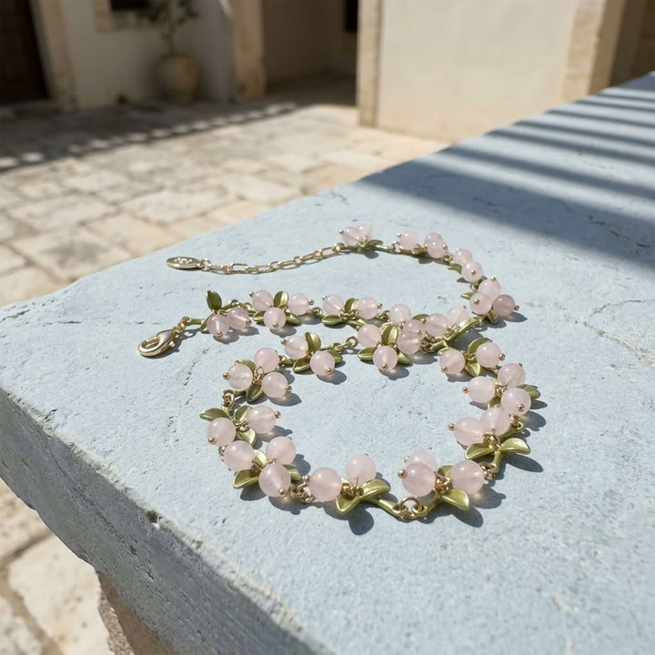 Rose Quartz Berry Garden Necklace with hand-painted green enamel leaves and gold-plated details displayed on a blue outdoor surface in sunlight.