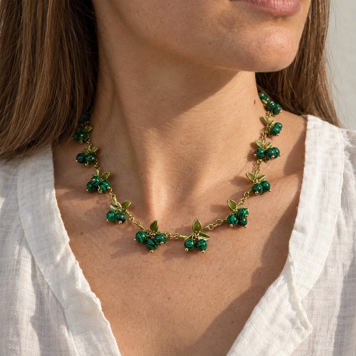 A close-up of a woman wearing a Malachite Berry Garden Necklace with natural green gemstone clusters and enamel leaf details, paired with a white blouse.