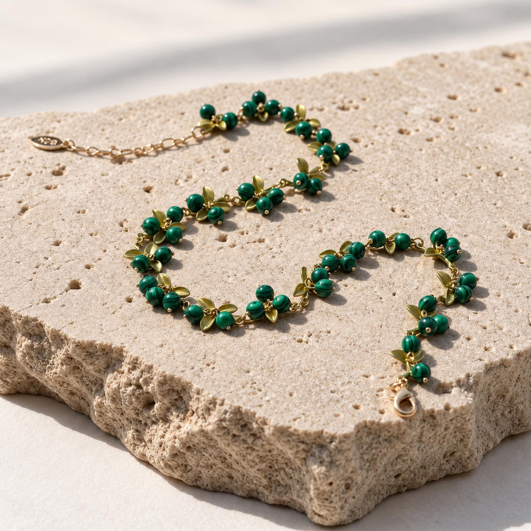 Malachite Berry Garden Necklace with striped green gemstone clusters, hand-painted enamel leaves and gold-plated details, arranged on a textured beige stone slab in bright natural light.
