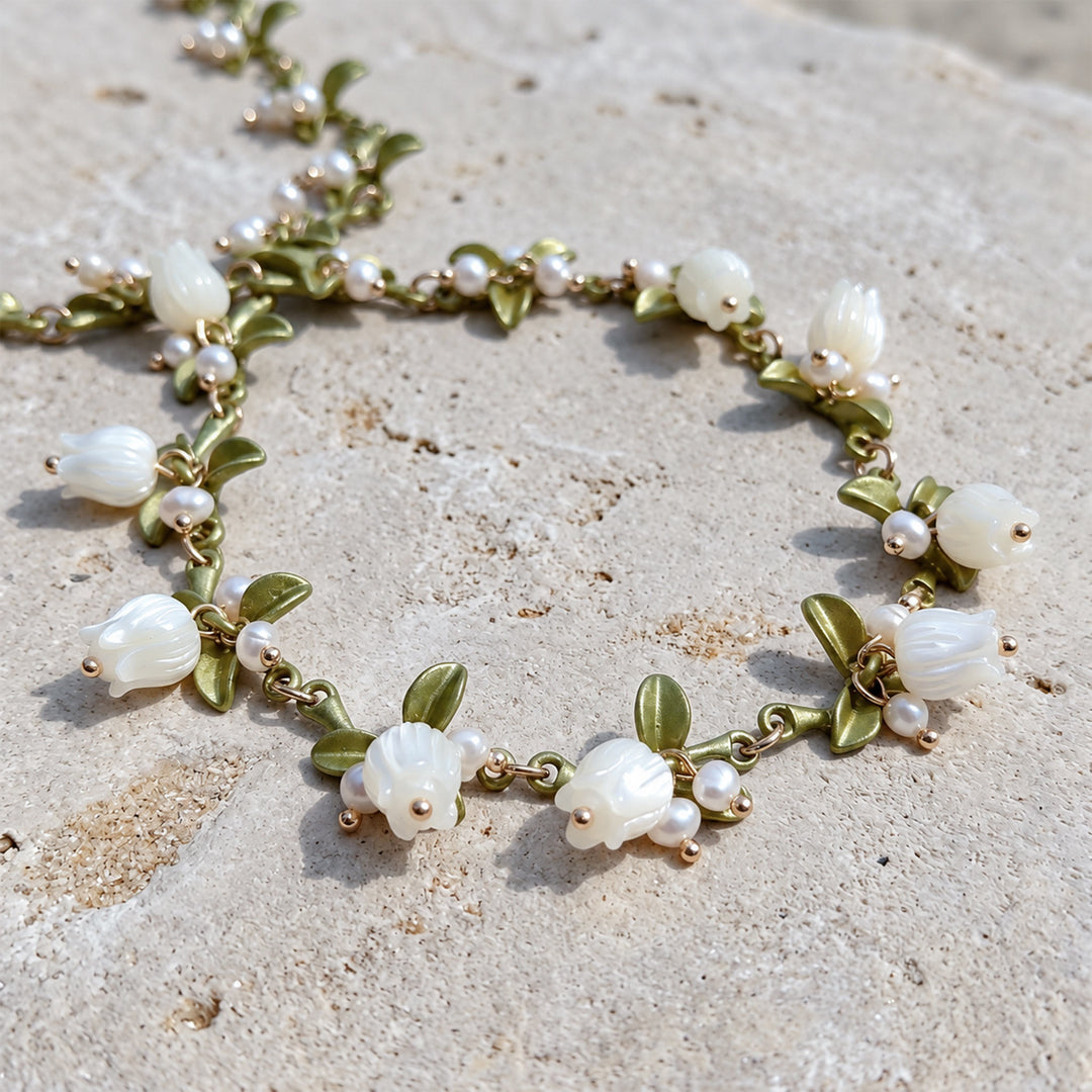Close-up of a MOP Lily of the Valley necklace with hand-carved mother-of-pearl flowers, freshwater pearls and green enamel leaves on stone surface.