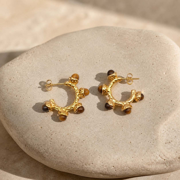 Gold hoop earrings with tiger eye stones on a stone surface