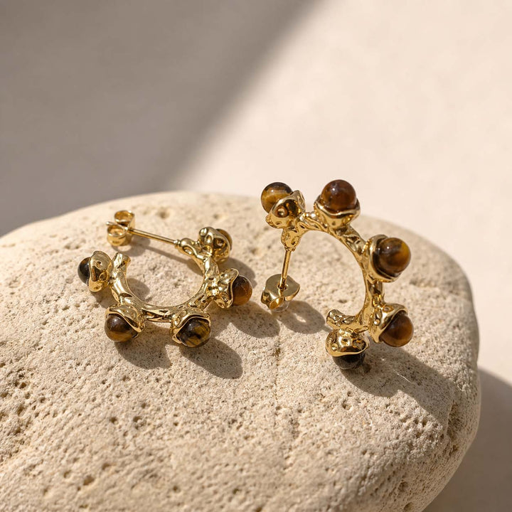 Gold hoop earrings withtiger eye gemstones on a textured stone surface