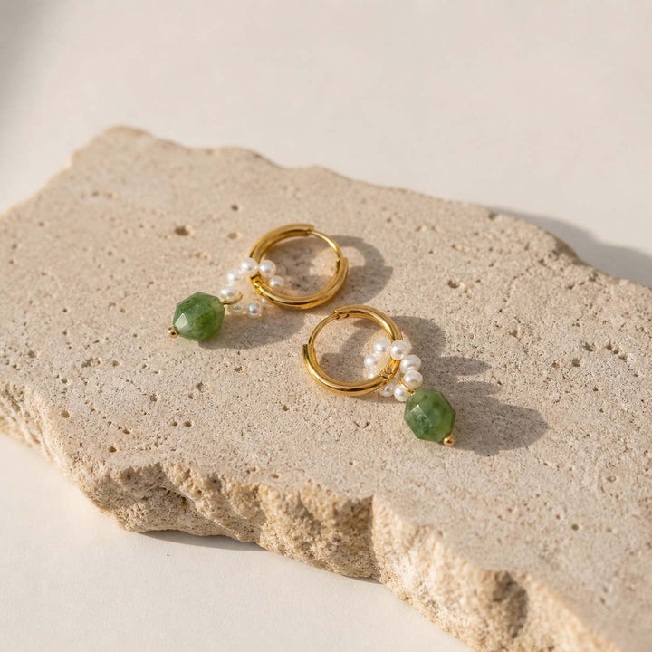Gold hoop earrings with pearl and green gemstone charms on sunlit stone
