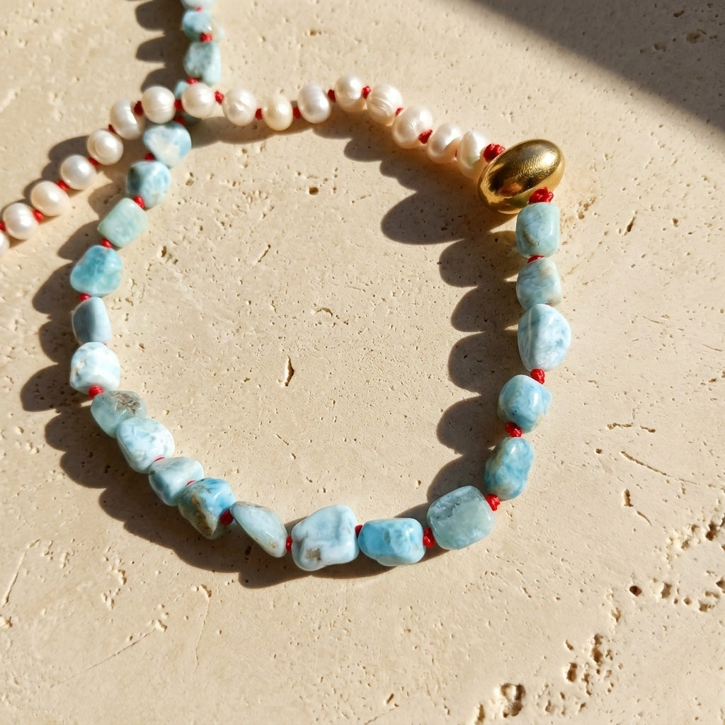 Close-up of Larimar and freshwater pearl necklace with red hand-knotted details and gold PVD stainless steel accent, displayed on a textured beige stone surface.