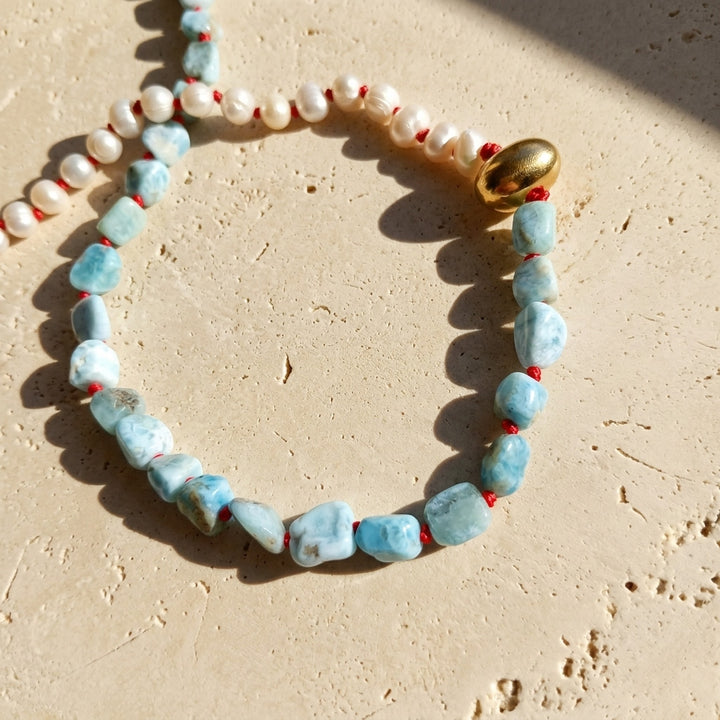 Close-up of Larimar and freshwater pearl necklace with red hand-knotted details and gold PVD stainless steel accent, displayed on a textured beige stone surface.