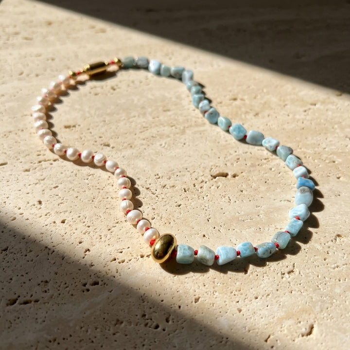Larimar pearl necklace arranged on a light stone surface with natural blue gemstone beads and gold magnetic clasp, displayed on a textured beige stone surface in direct sunlight.