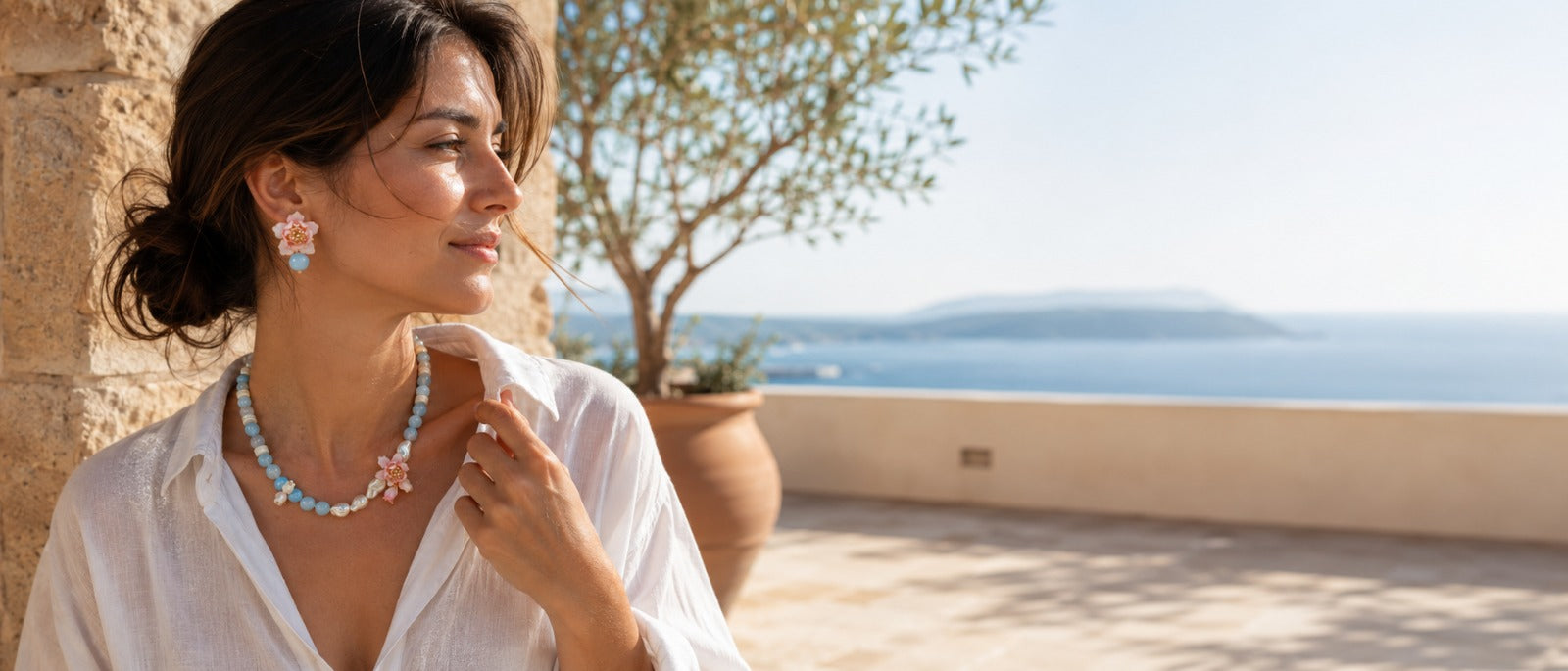 Woman wearing A2K London floral earrings and an aquamarine pearl necklace on a sunlit terrace by the sea